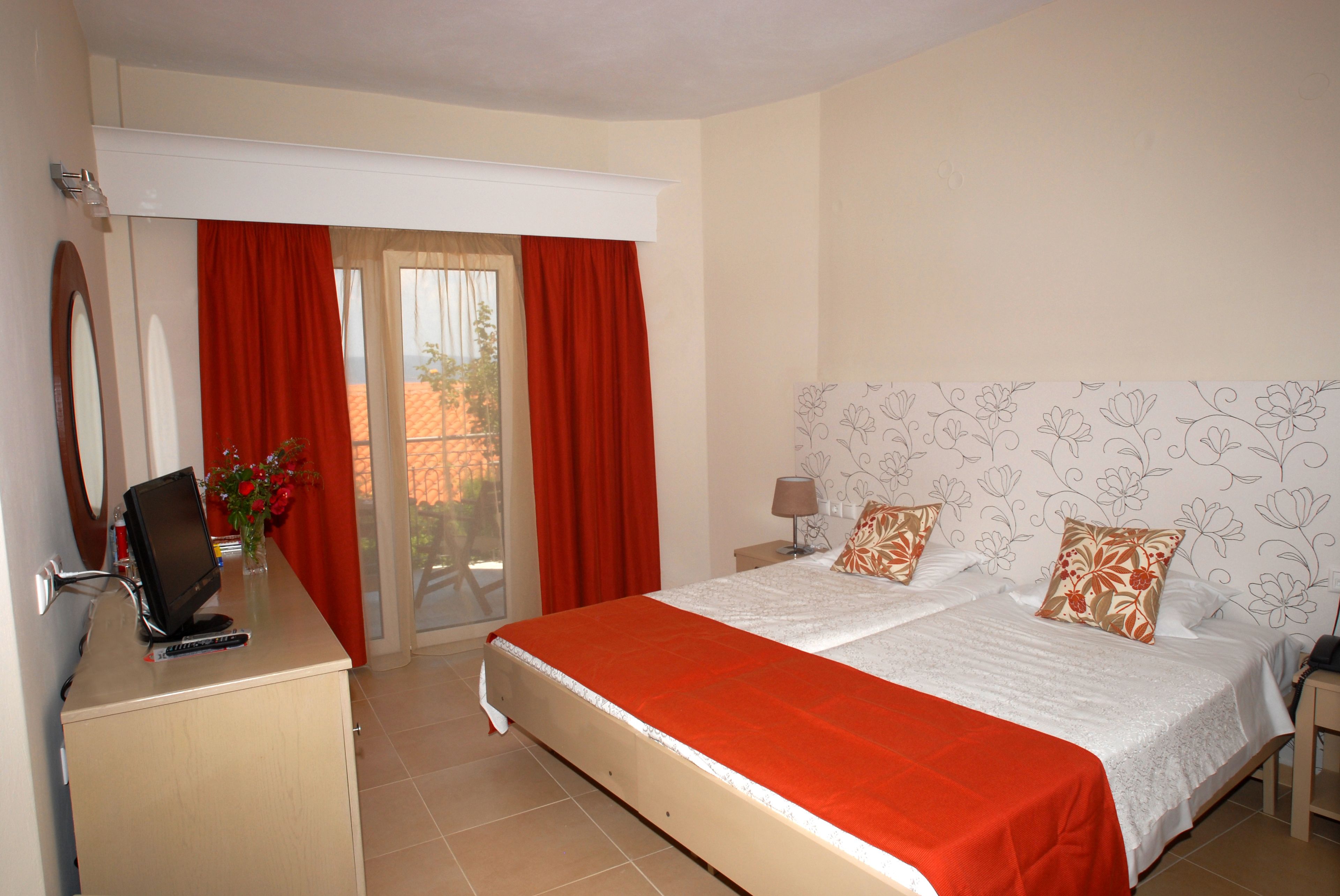 A hotel room with a bed, a desk with a computer, a chair, and a window with red curtains.