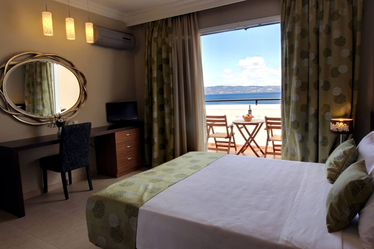 A hotel room with a bed, a desk with a television, a chair, a small table with a fruit bowl, and a view of the sea.