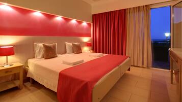Een hotelkamer met een bed, nachtkastjes, een rood hoofdeinde, een balkon met gordijnen en een uitzicht op de stad in de schemering.