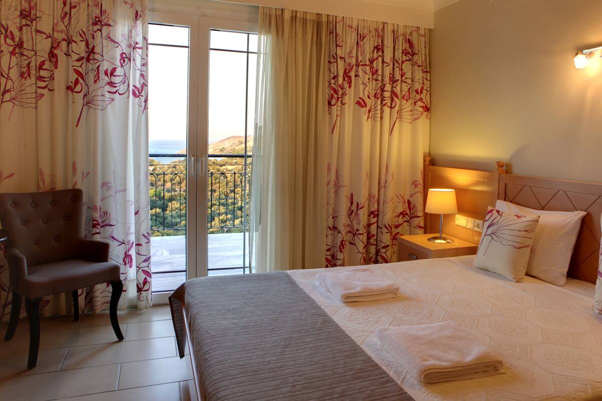 A hotel room with a bed, a chair, a nightstand with a lamp, and a view of a balcony with curtains.