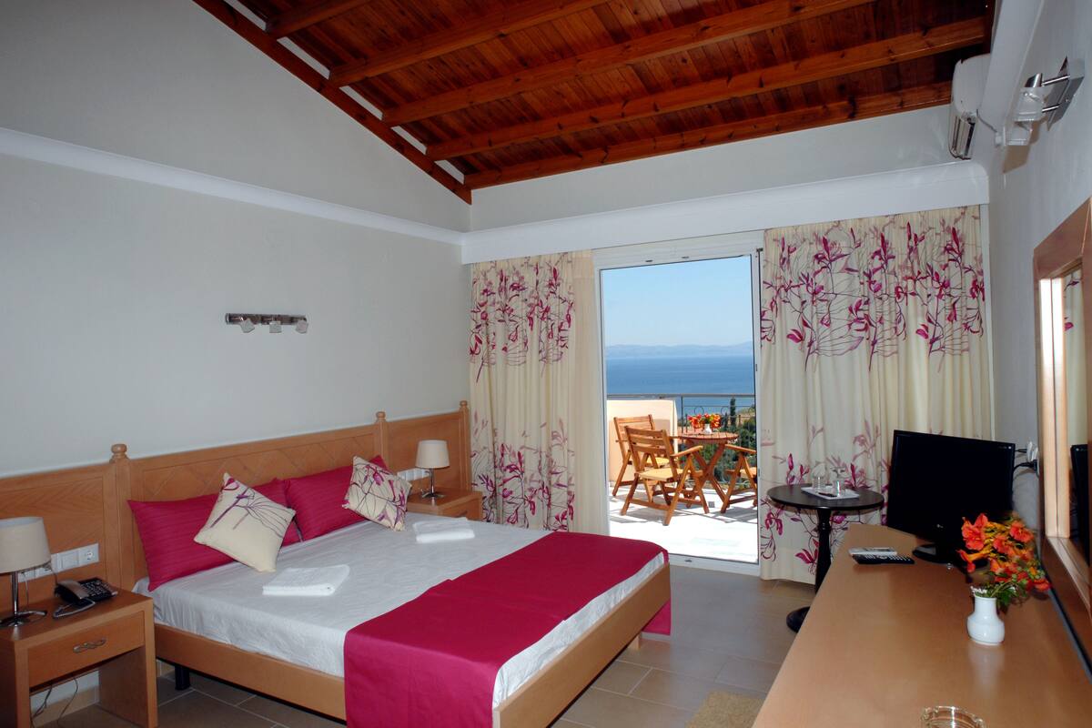 A hotel room with a wooden ceiling, a bed with a red bedspread, a desk with a TV, and a view of the sea through the window.