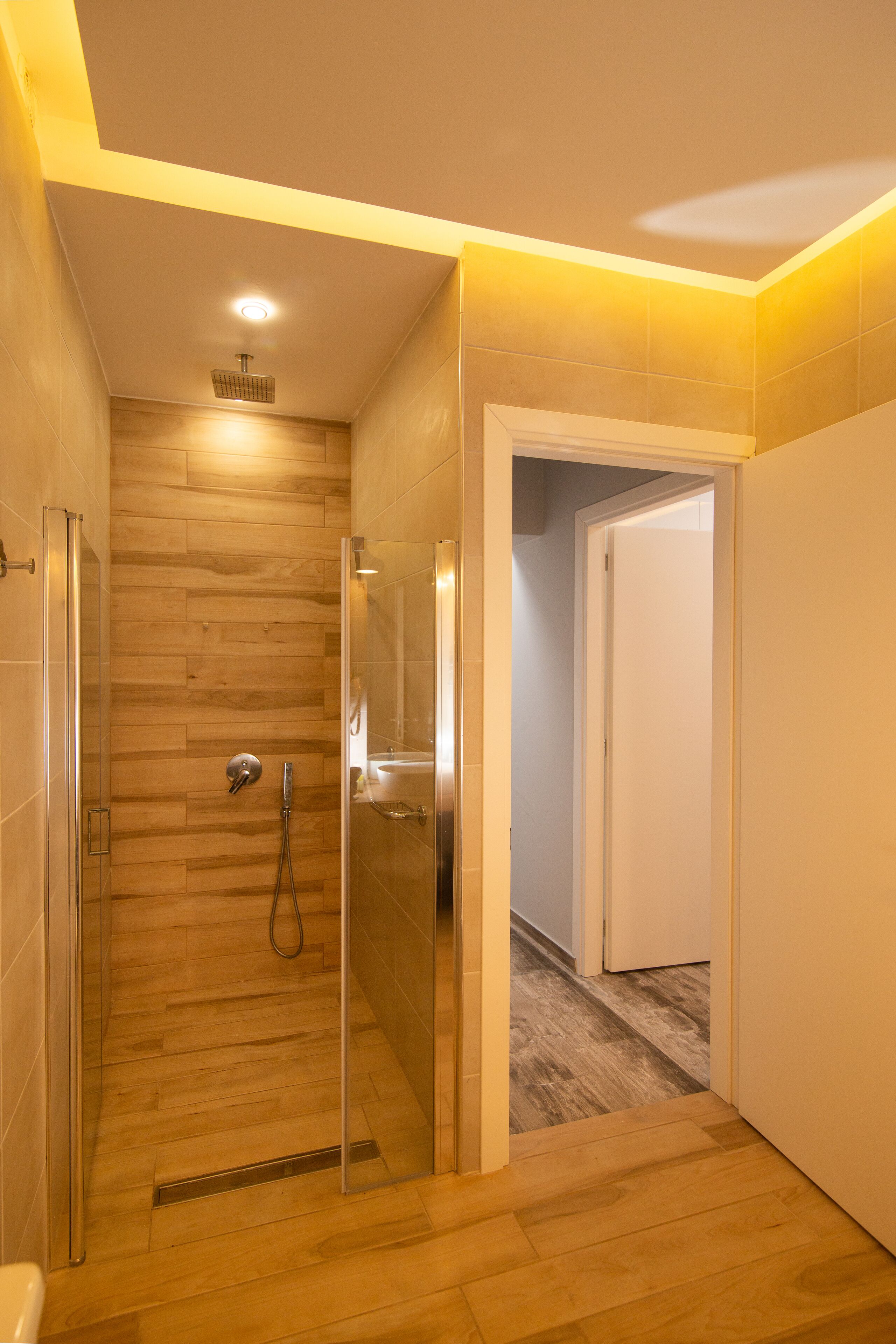 Un baño moderno con ducha envuelta en cristal, suelo de madera y una puerta blanca que da a otra habitación.