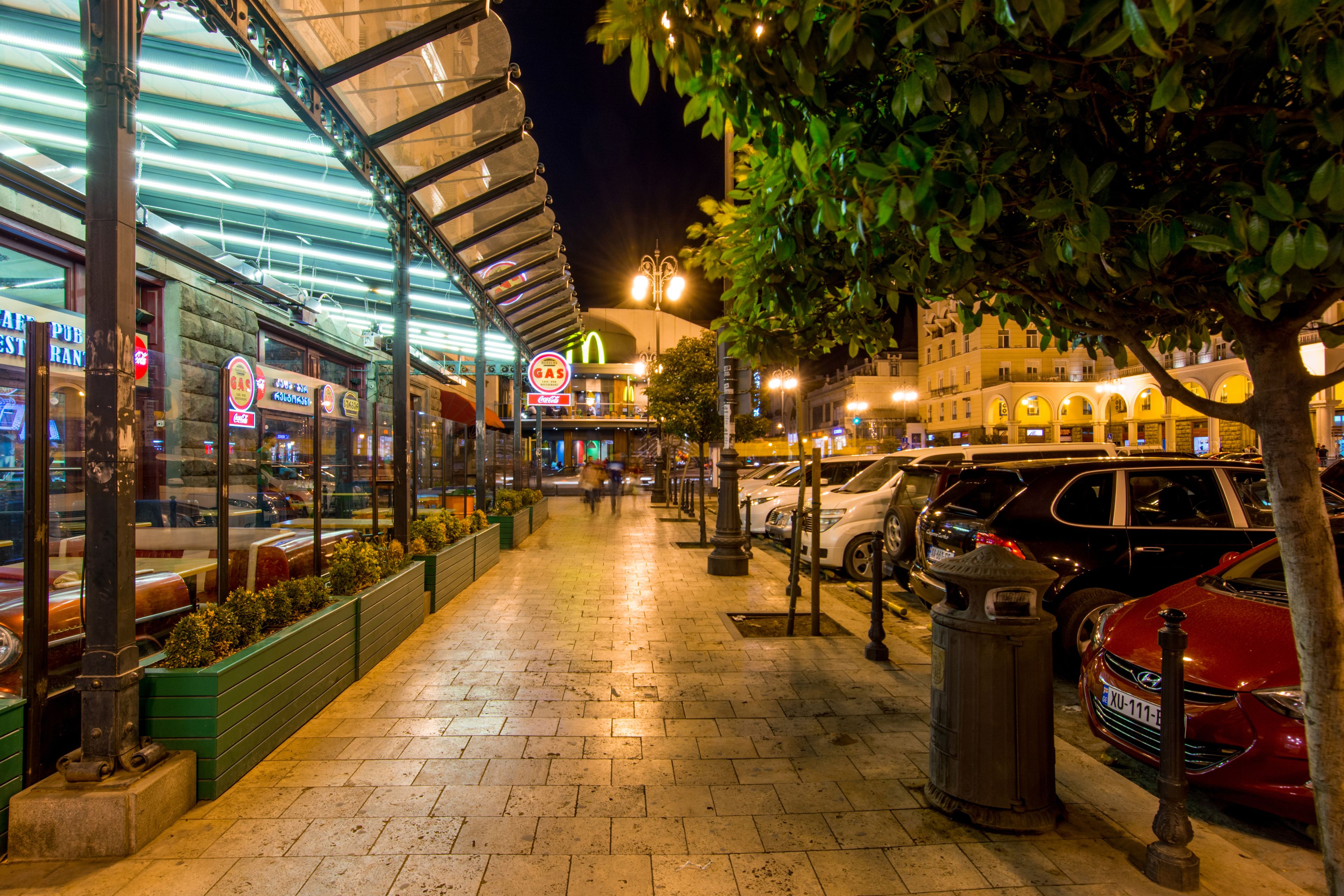A nighttime urban street scene with a restaurant, parked cars, and a McDonald's.