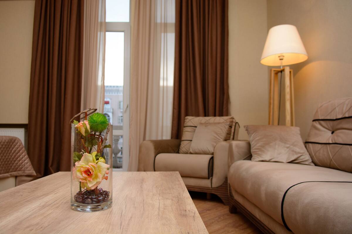 A room with a wooden table, a vase with flowers, a sofa, and a lamp.