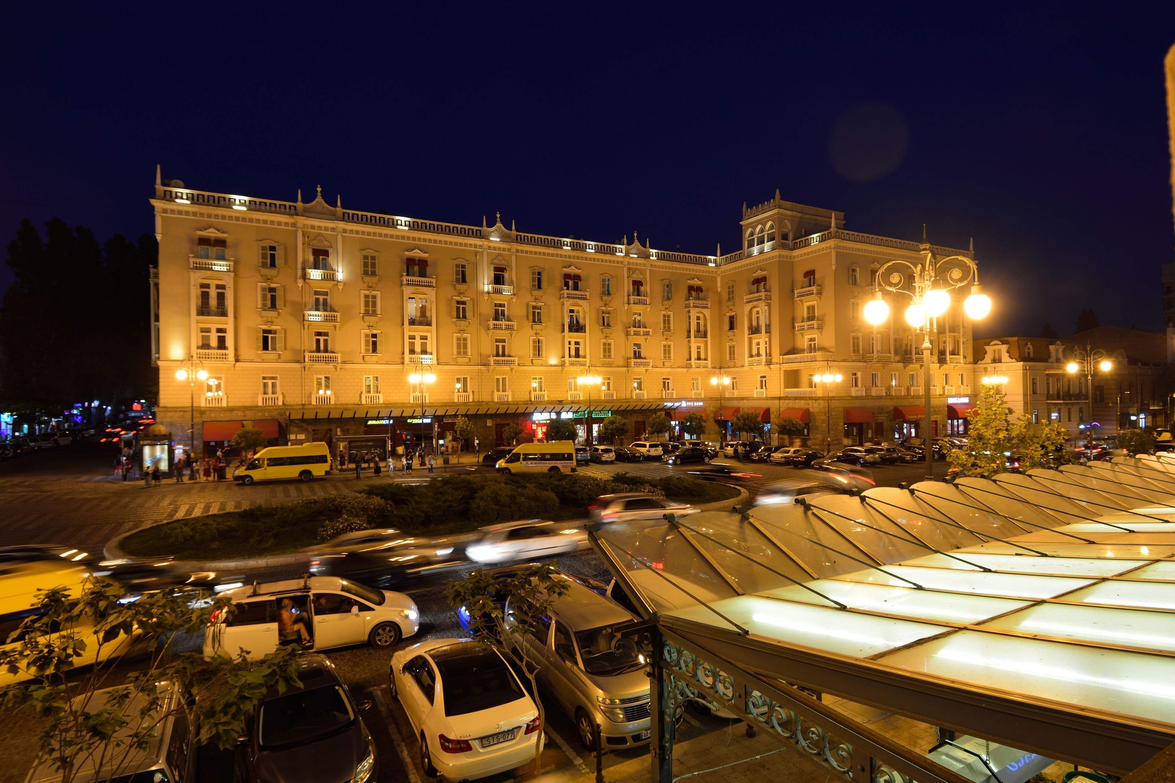 Un grand hôtel illuminé, doté de nombreux étages et de balcons, un parking avec plusieurs voitures et un espace extérieur couvert aménagé en coin salon.