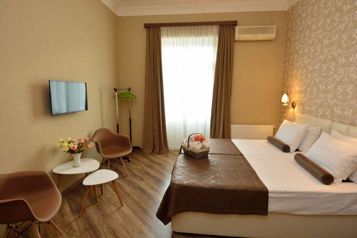 A hotel room with a bed, two chairs, a small table with a vase of flowers, a standing fan, and a television mounted on the wall.