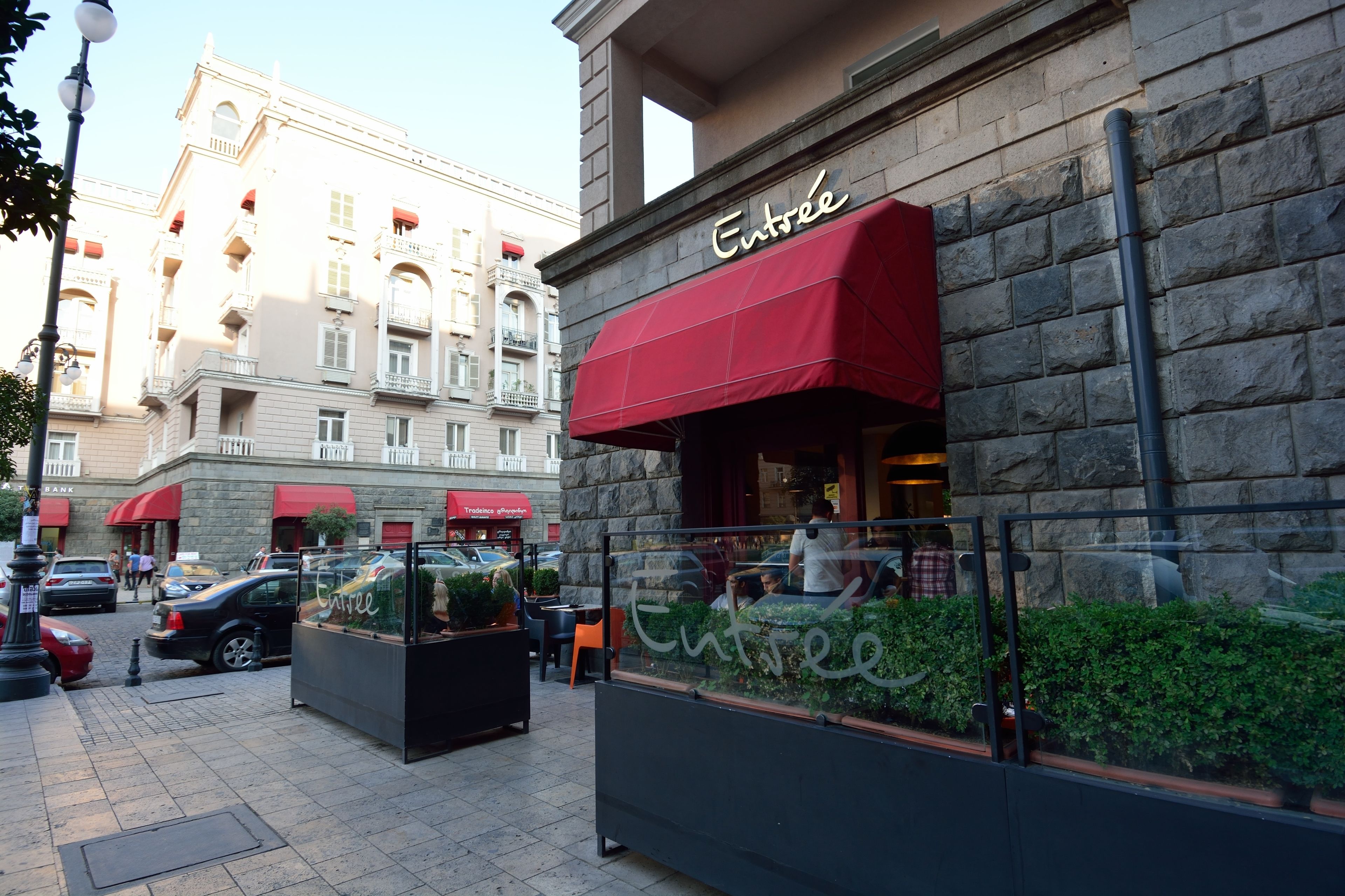 A building with a red awning labeled "Entrée" and a black planter with greenery in front.