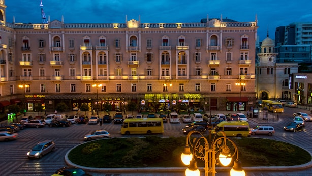 Un edificio grande e histórico con varios pisos y balcones, rodeado por una calle urbana concurrida con coches y un autobús amarillo.
