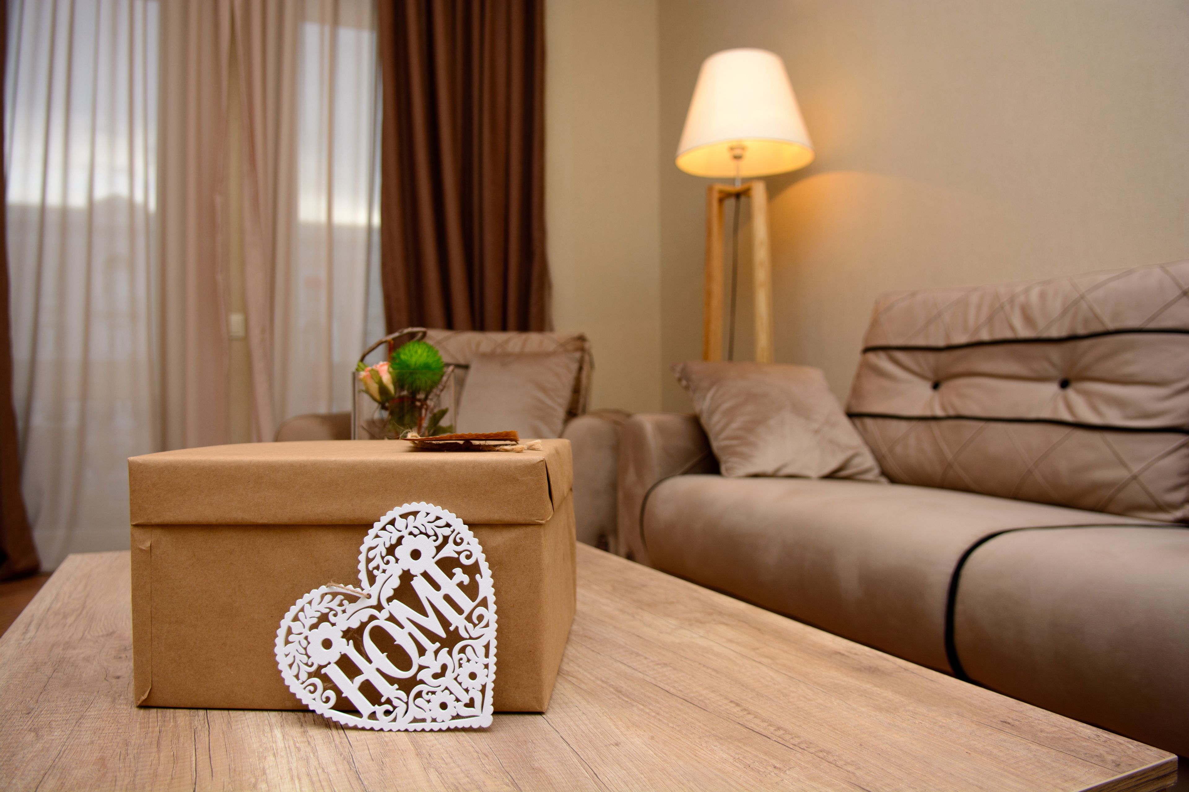 Una caja de regalo envuelta en papel marrón con un adorno blanco en forma de corazón sobre una mesa de madera en una sala de estar con un sofá beige y una lámpara de pie.