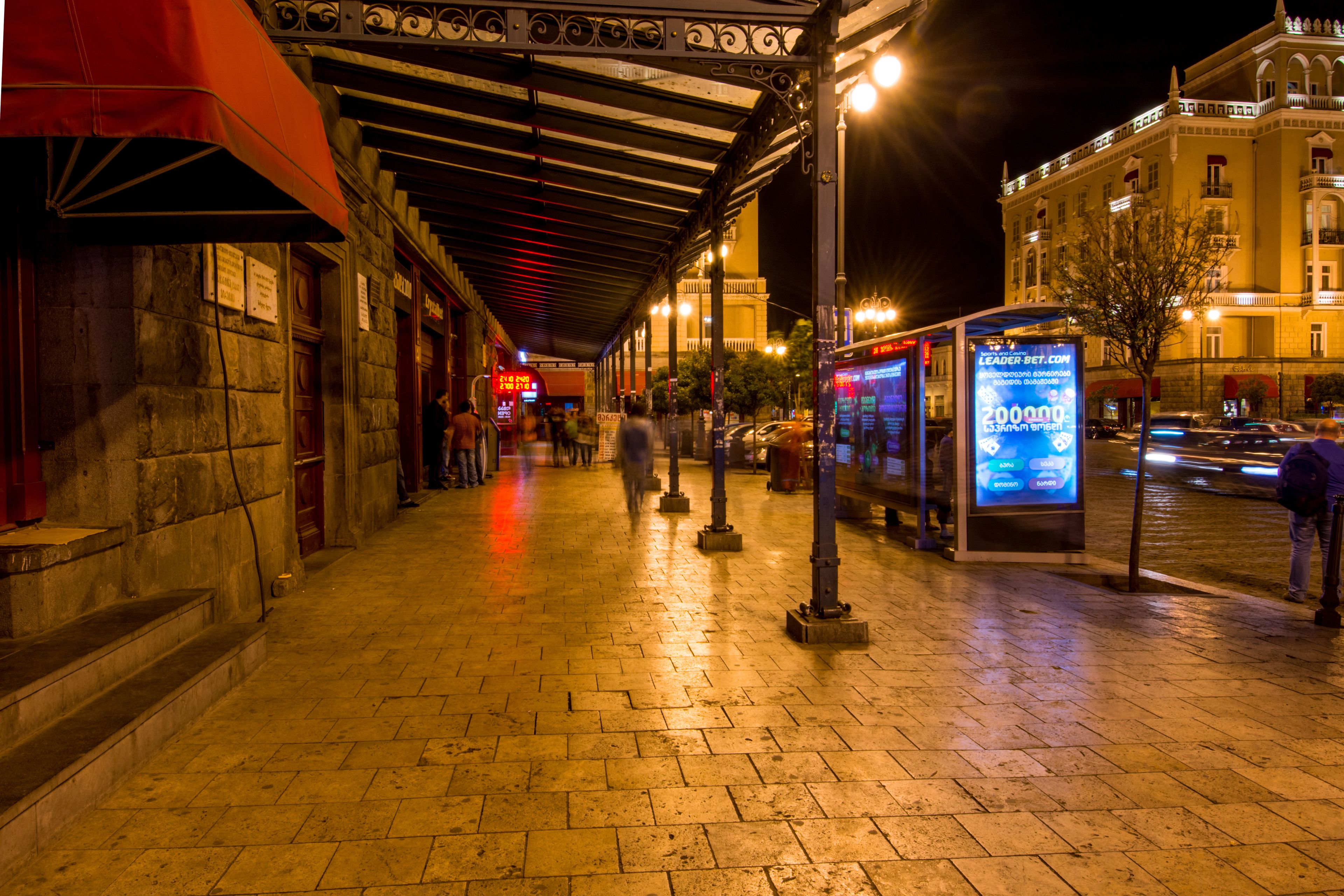 A nighttime urban street scene with a covered walkway, stone buildings, and a digital advertisement board.