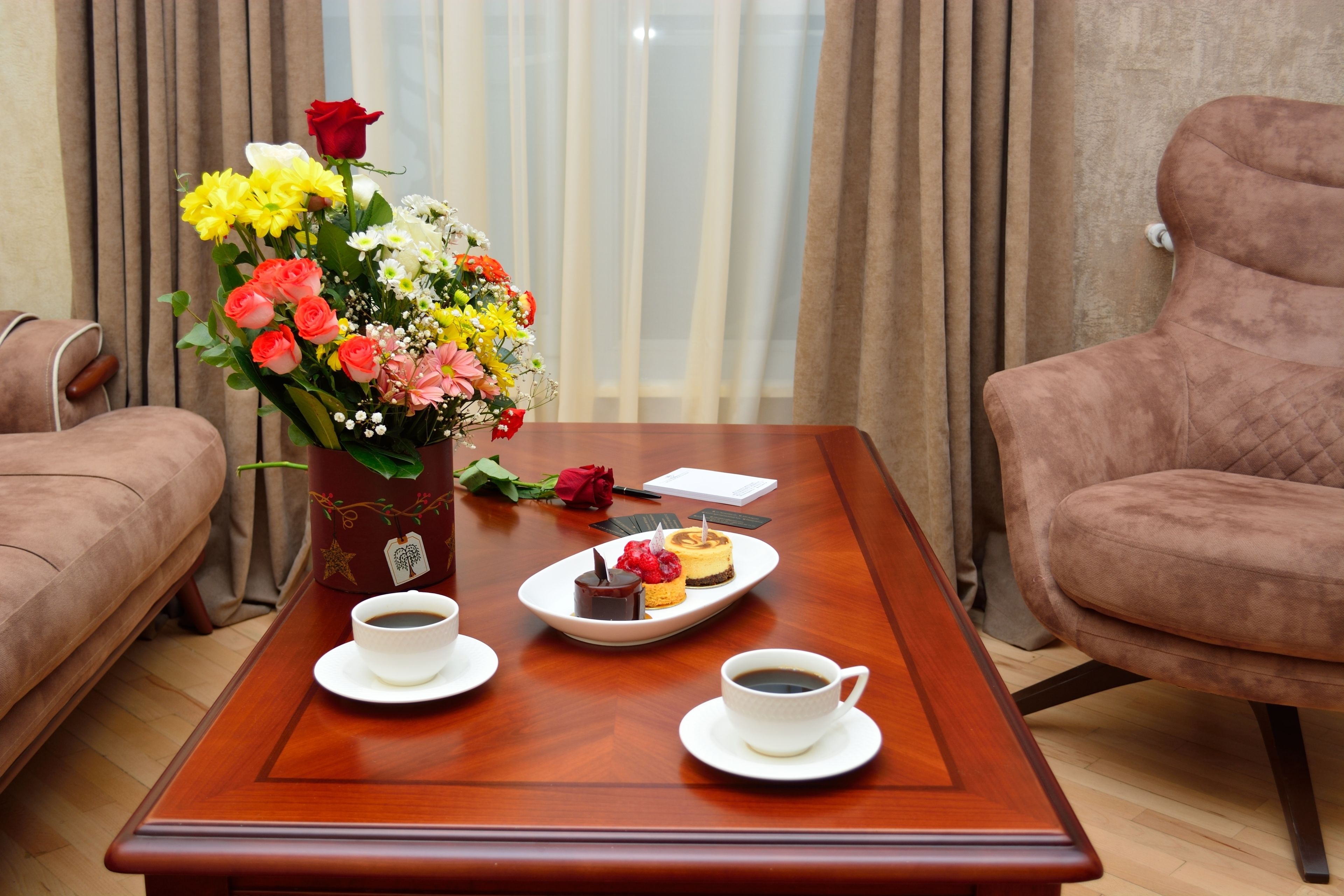 A wooden coffee table with two cups of coffee, a plate with desserts, and a bouquet of flowers.