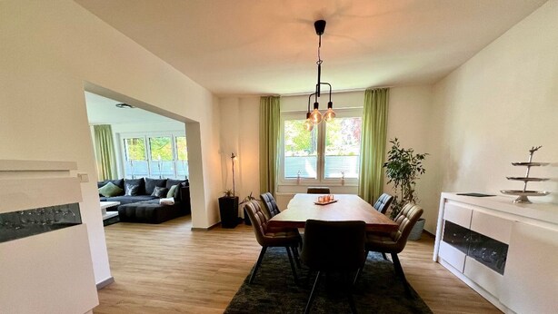 A modern kitchen with a dining table, chairs, and a chandelier. There is a sofa and a plant in the background. - Pension Alscher (Goslar)