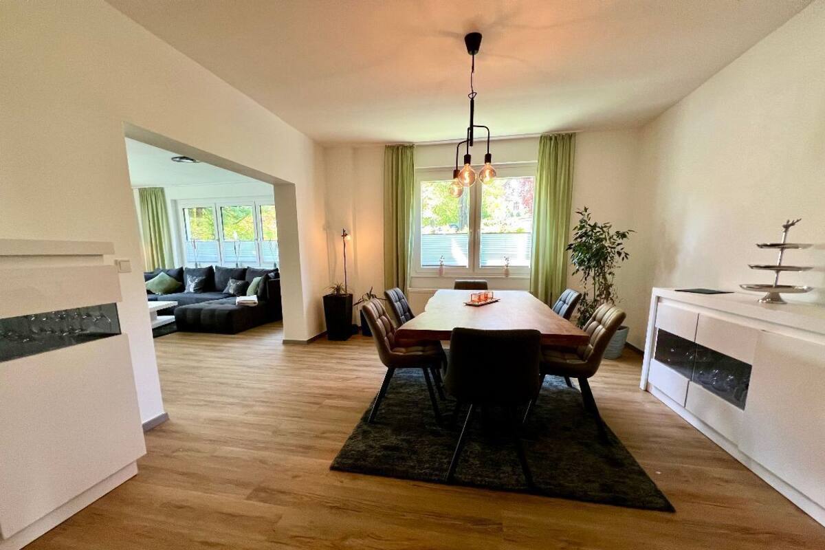 A modern kitchen with a dining table, chairs, and a chandelier. There is a sofa and a plant in the background.