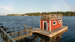Un muelle de madera que lleva a un pequeño edificio rojo con un flotador y una puerta, rodeado de agua tranquila y una orilla boscosa.