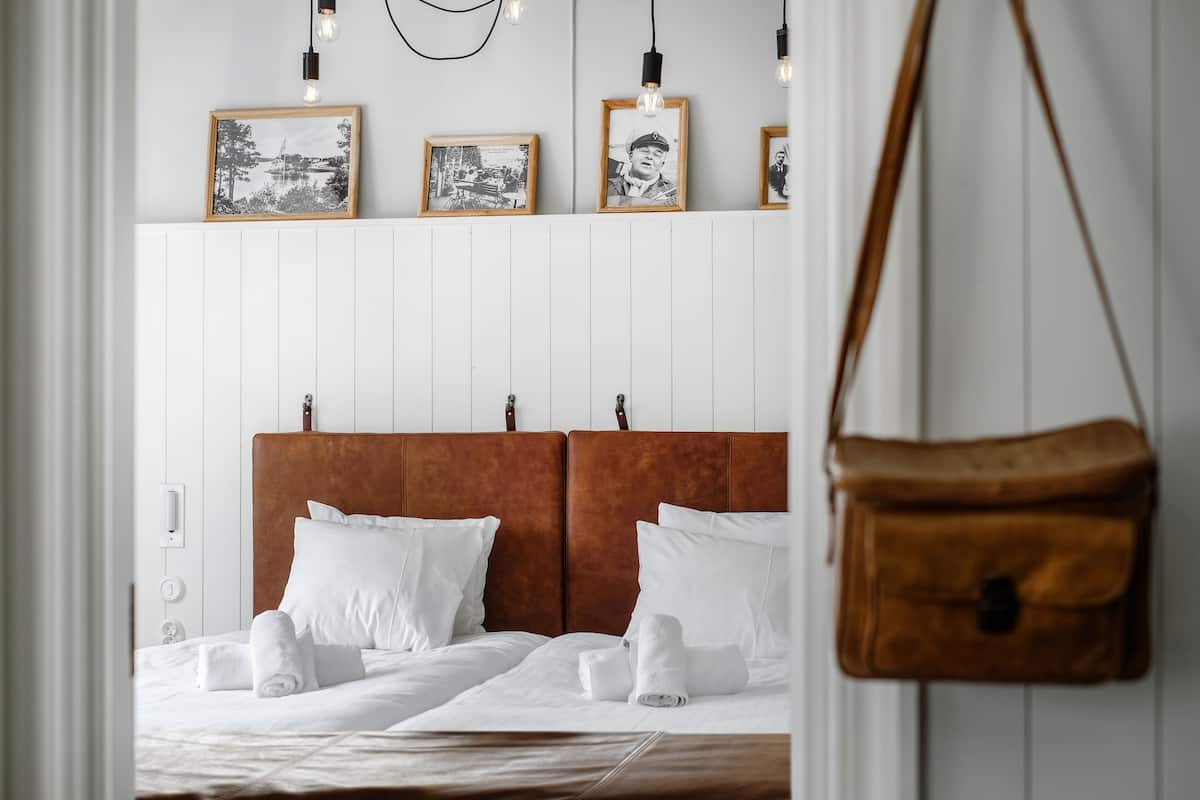 A hotel room with a white bed, two pillows, a brown headboard, and a hanging brown bag.