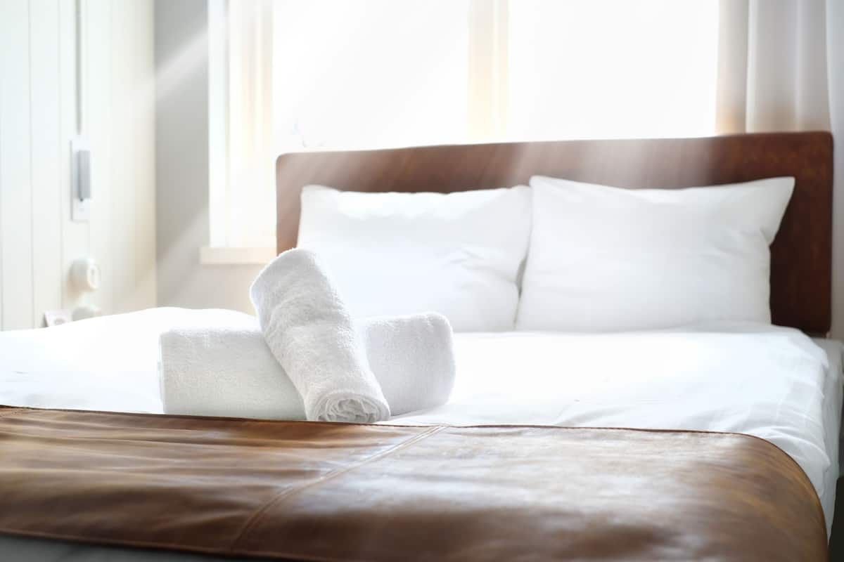 A neatly made bed with white linens and a brown leather ottoman.