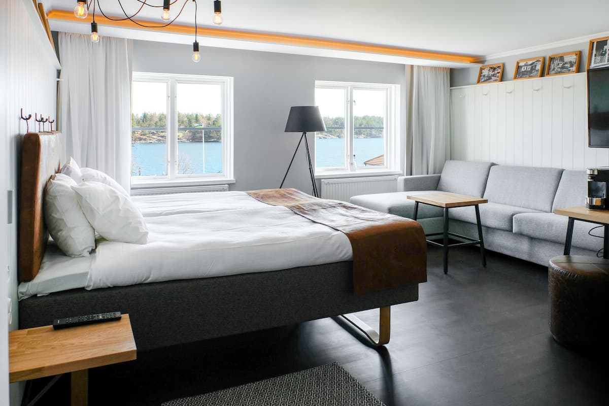A modern hotel room with a bed, a sofa, a small table, and a TV. There are windows with a view of a body of water and a lamp.