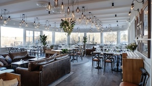 Un restaurante espacioso con techos altos, vigas a la vista y numerosas lámparas colgantes. Las mesas están preparadas con copas de vino y platos, y hay plantas en macetas y un sofá grande en la zona de estar.