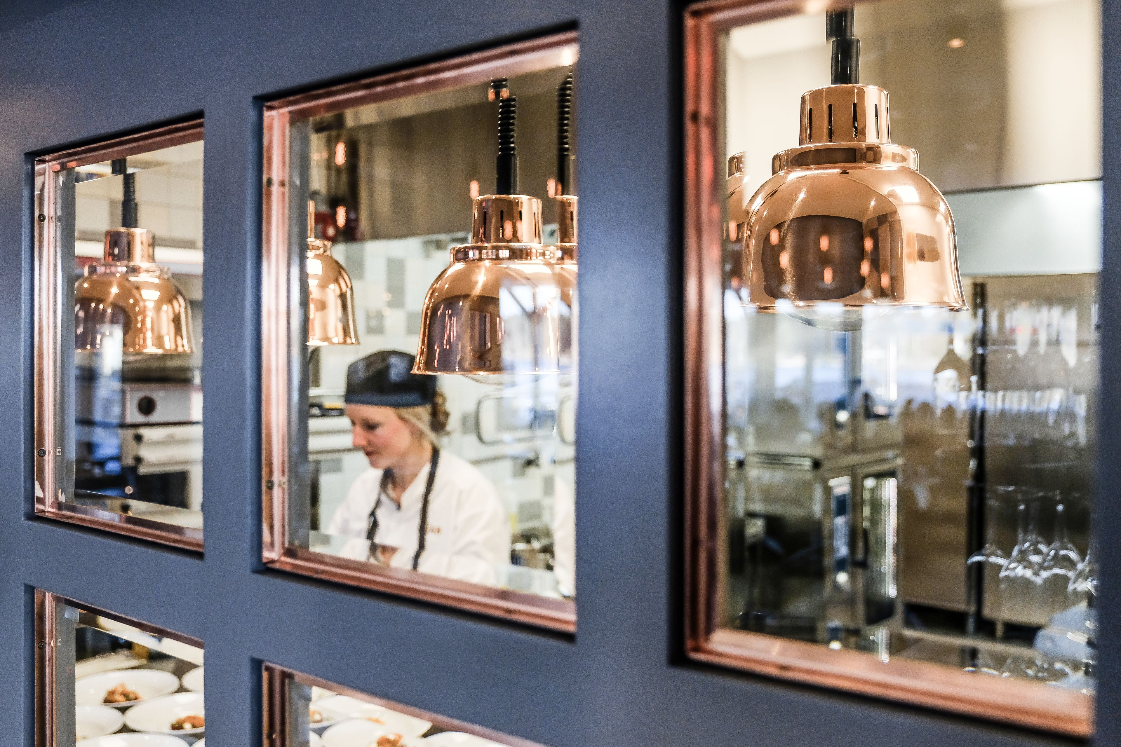 A wall with multiple framed mirrors reflecting a kitchen area with copper pendant lights and a chef in uniform.