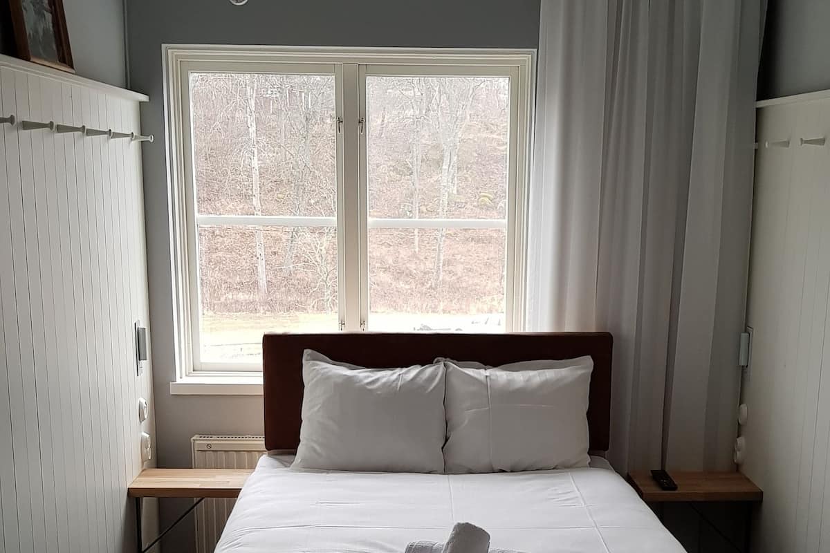 A neatly made bed with white linens and a folded towel, a bedside table with a lamp, a window with curtains, and a patterned rug on the floor.