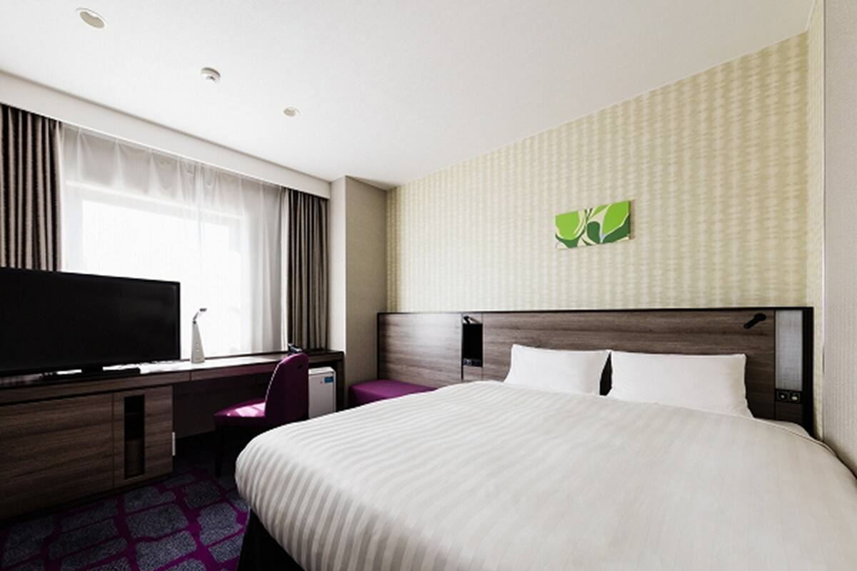 A hotel room with a large bed, a desk with a flat-screen TV, a chair, and a small table.