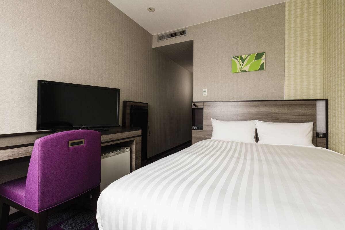 A hotel room with a bed, a desk with a television, and a purple chair.