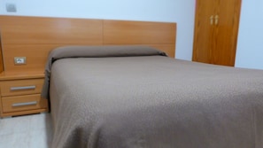 Un dormitorio con cabecera de madera, una cama individual con colcha color beige y una mesita de noche con cajones.