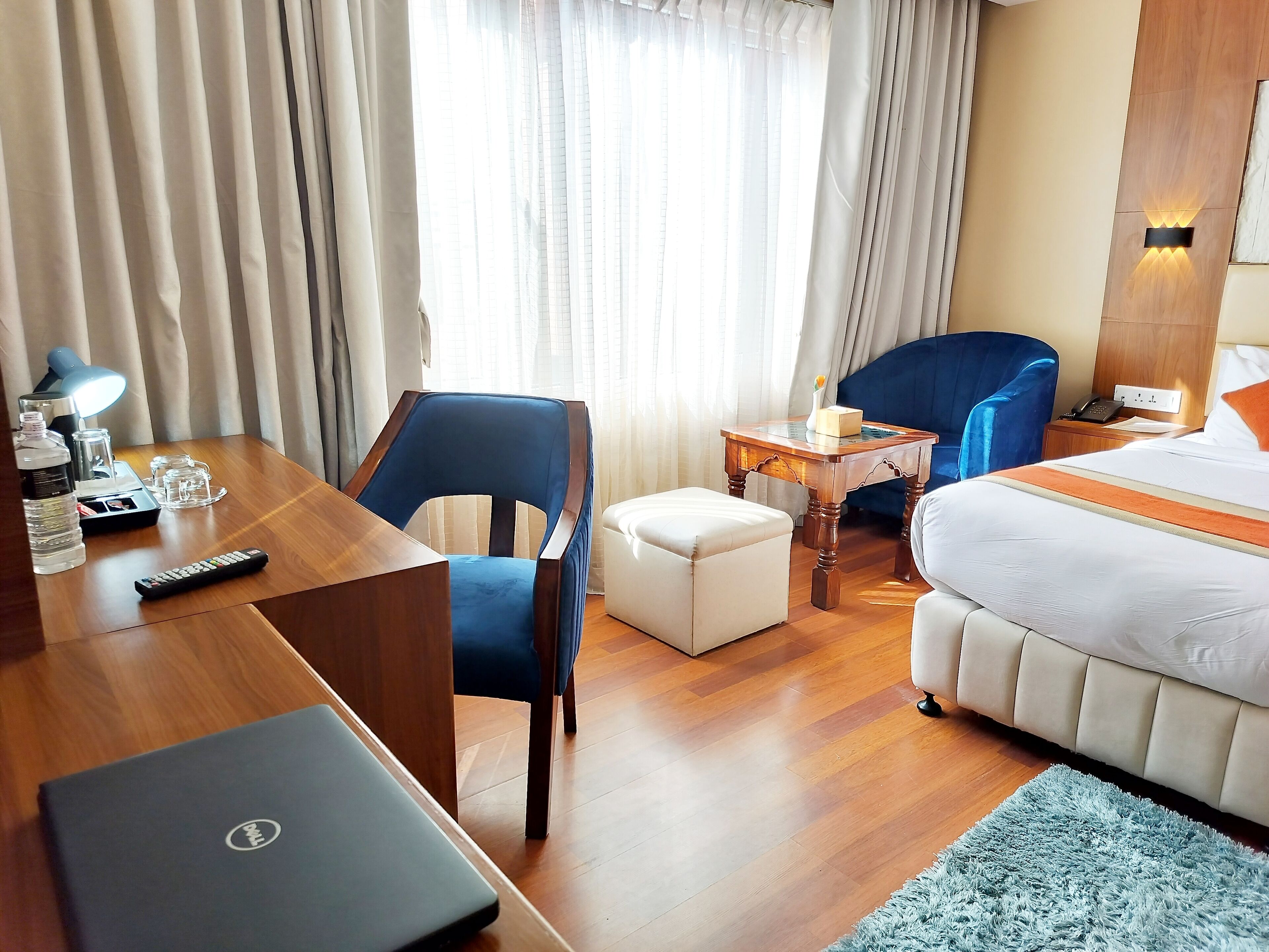 Una habitación de hotel con un escritorio de madera, una silla azul, una mesita con una lámpara, una cama con una colcha blanca y una alfombra azul.
