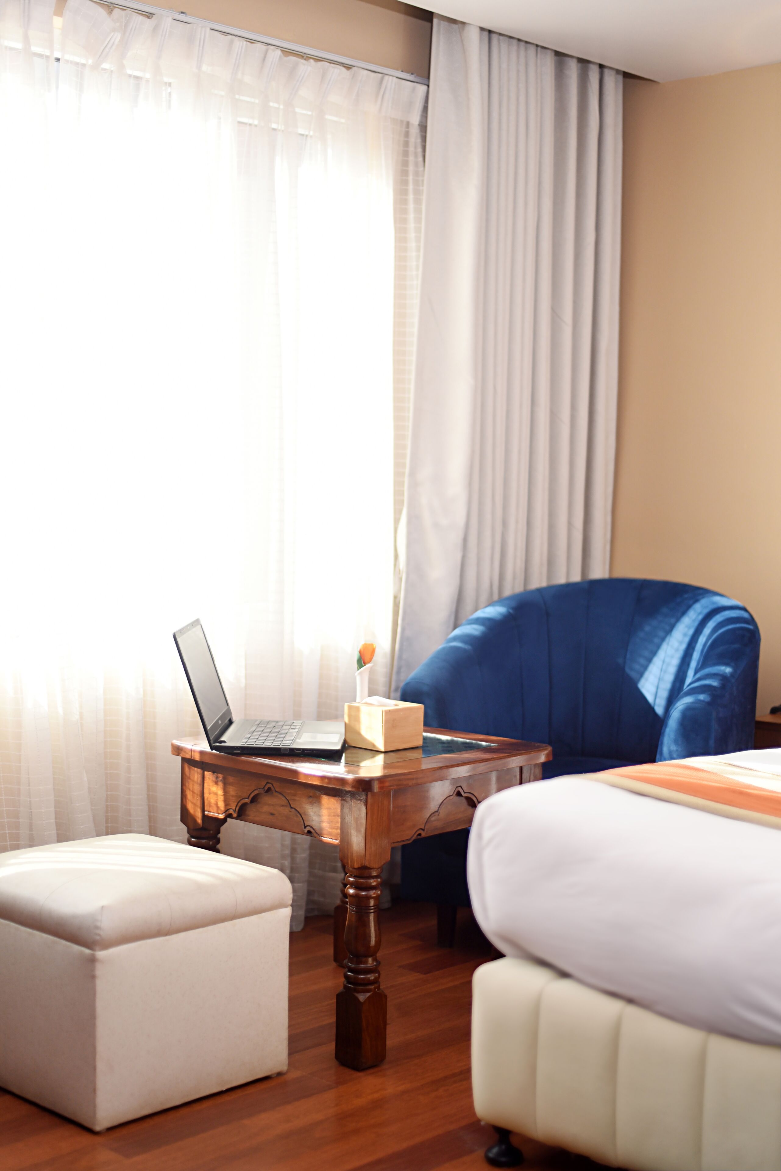 Une chambre d’hôtel avec un ordinateur portable posé sur une petite table en bois, une chaise bleue, un pouf blanc et un lit avec une literie blanche.
