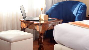 Une chambre d’hôtel avec un ordinateur portable posé sur une petite table en bois, une chaise bleue, un pouf blanc et un lit avec une literie blanche.