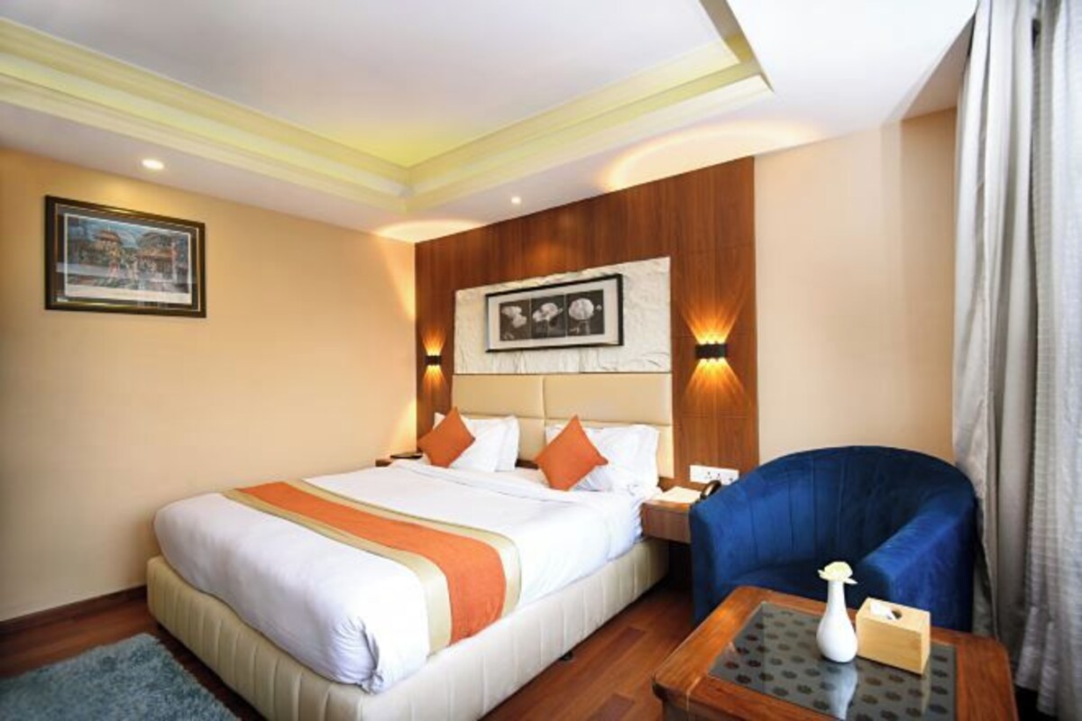 Een hotelkamer met een groot bed, een blauwe fauteuil, een klein tafeltje met een vaas, en lijstjes met foto's aan de muur.
