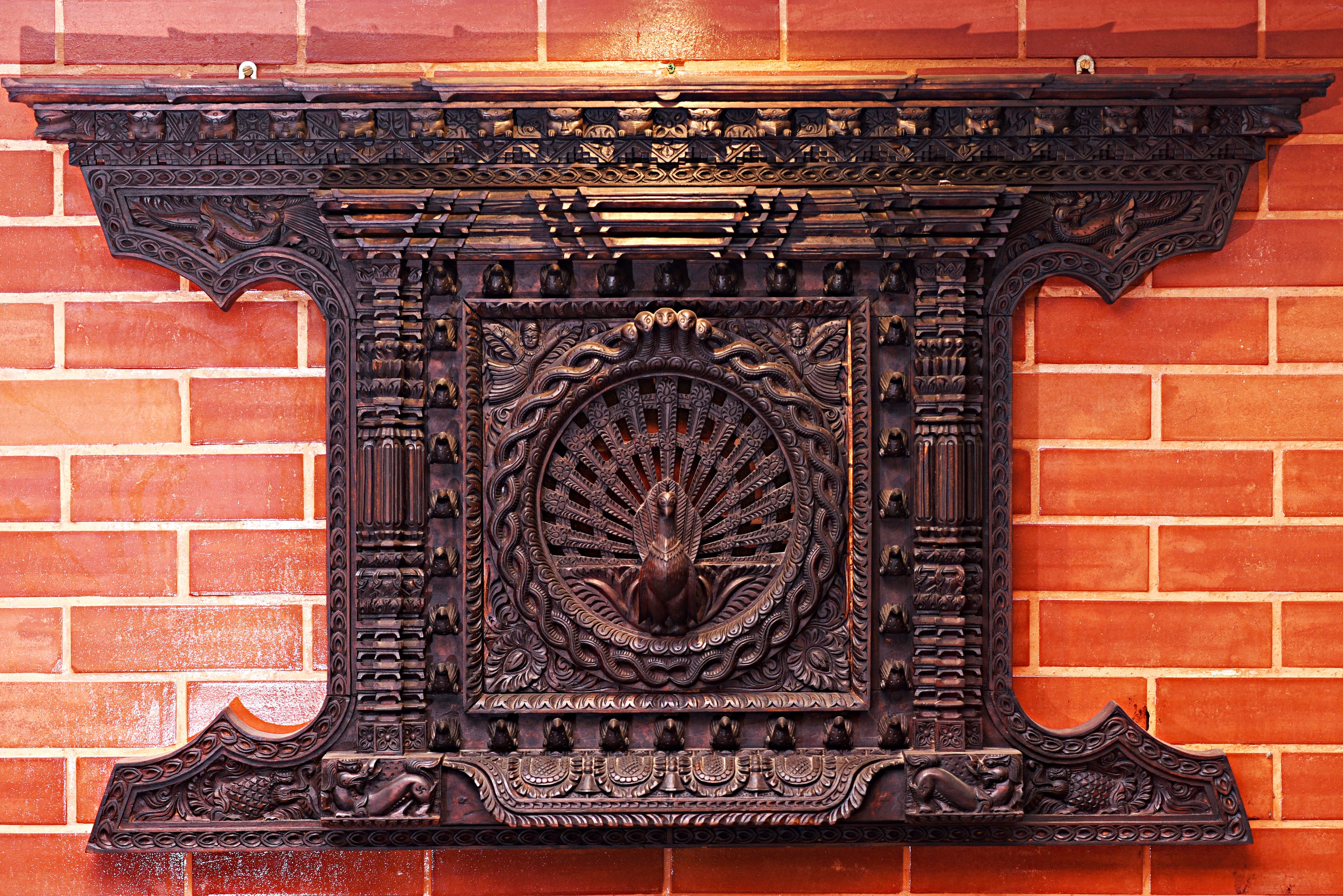 A detailed, ornate fireplace with intricate carvings and a central peacock motif, set against a brick wall.