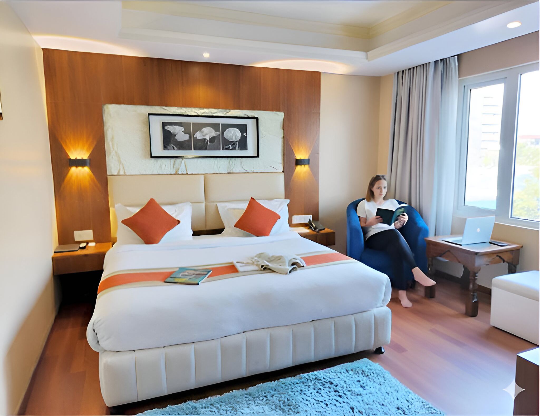 A hotel room with a large bed, a reading chair, a desk with a laptop, and a window with curtains.