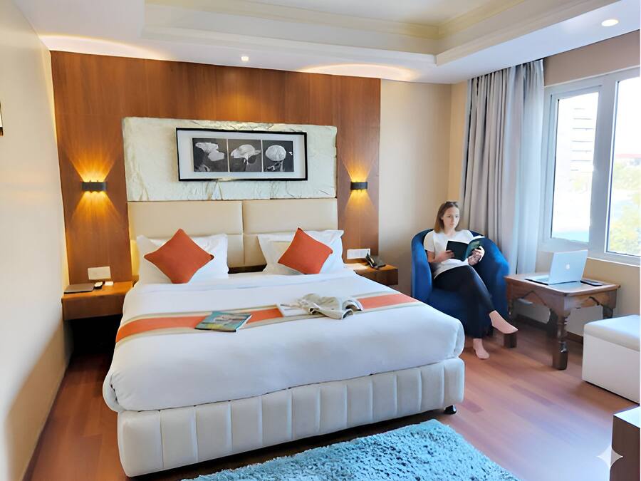A hotel room with a large bed, a reading chair, a desk with a laptop, and a window with curtains.