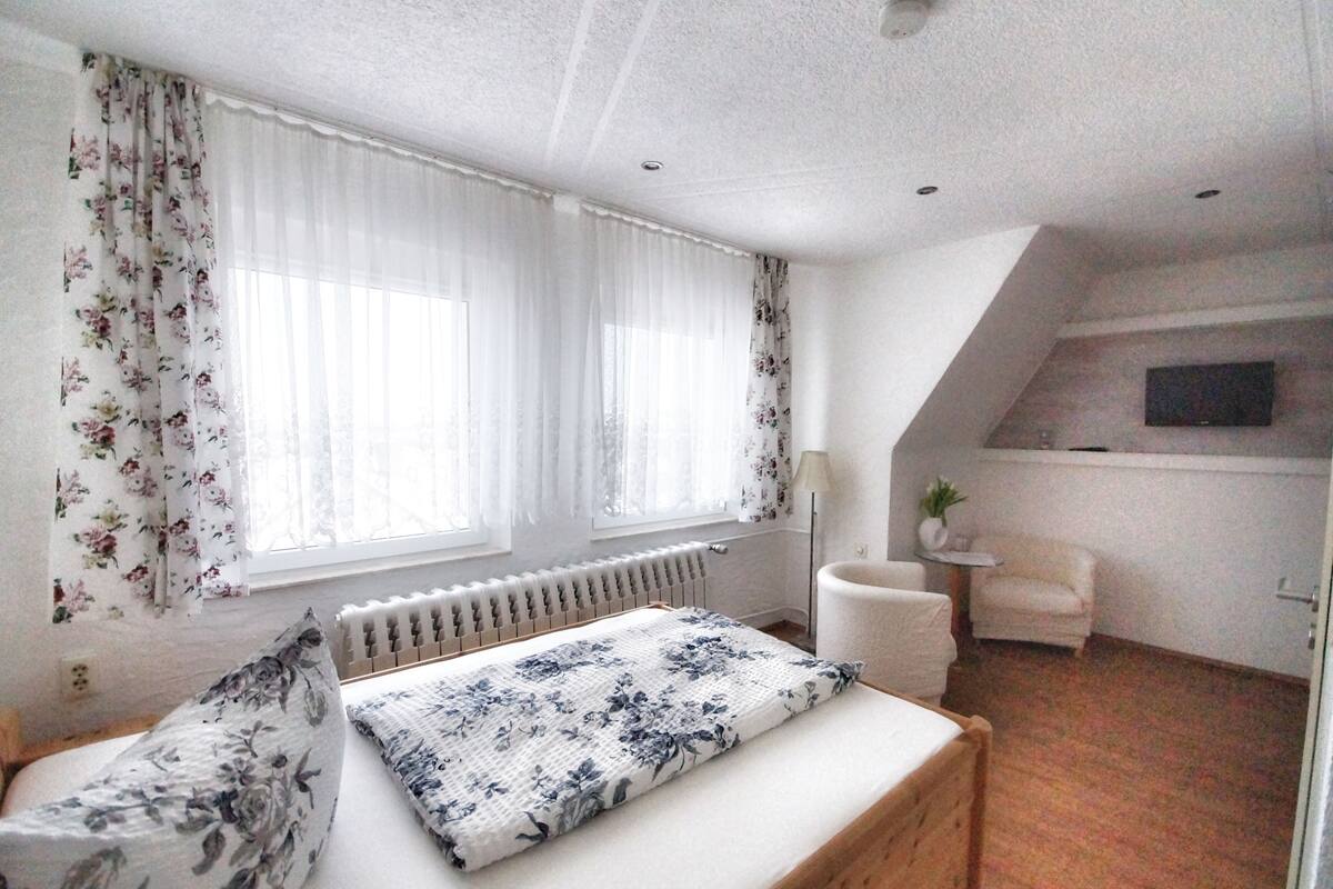 A bedroom with a bed, floral curtains, a radiator, a chair, and a television on the wall.
