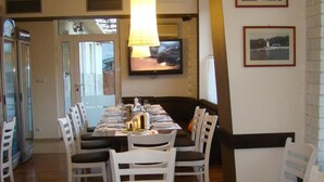 A dining area with a long table set for four, surrounded by white chairs. A large lamp hangs above the table, and a television is mounted on the wall. - Hotel Lilia 2 (Primorsko)