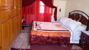 Un dormitorio con un armario de madera, una cama con colcha estampada, una cortina roja y una ventana pequeña.