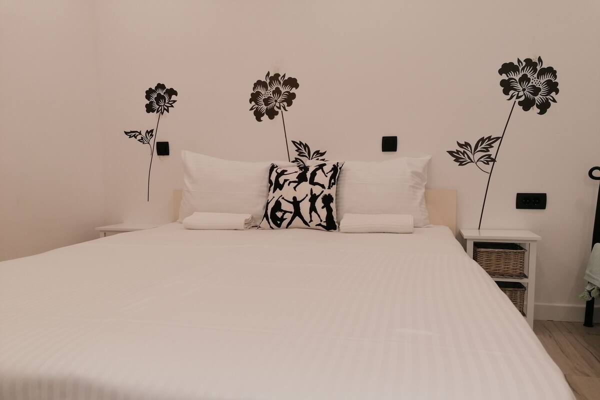 A neatly made bed with white linens and black floral-patterned pillows, a small bedside table with a basket, and a wall-mounted light switch.