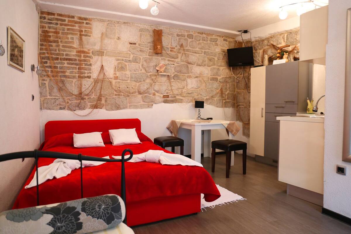 A compact room with a red bed, a small desk, a white cabinet, and a stone wall.