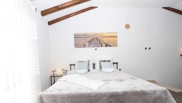 A neatly made bed with a white duvet and pillows, a framed picture on the wall, and a wooden ceiling beam.