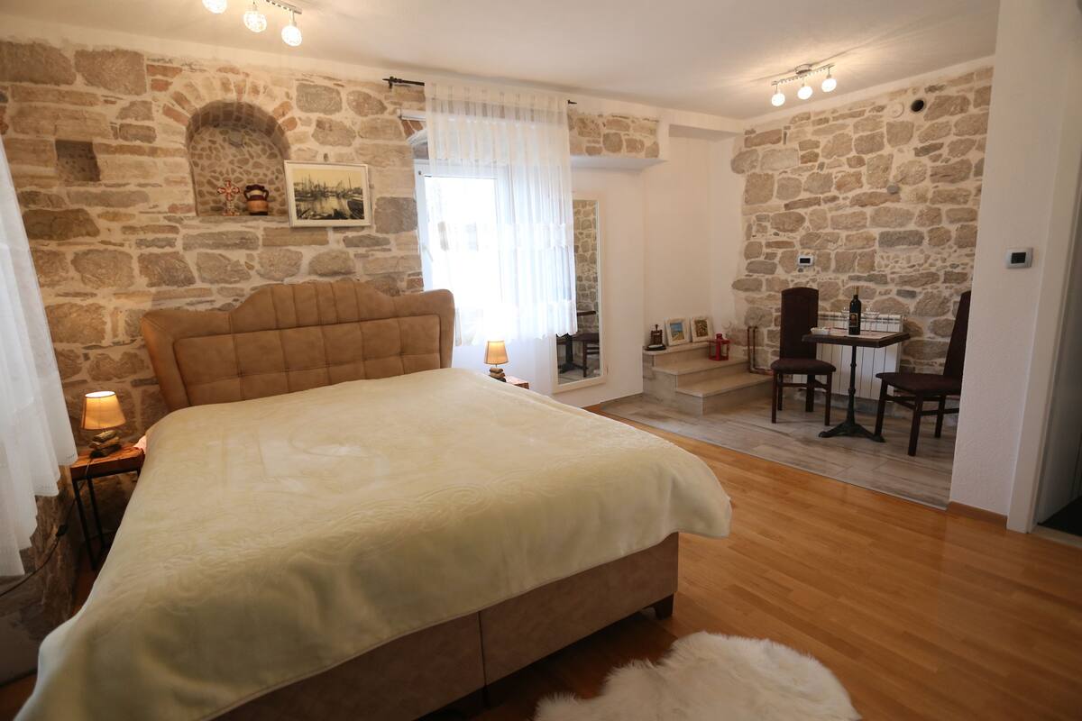 A bedroom with a stone wall, a bed with a headboard, a small table with chairs, and a window with curtains.
