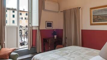Un dormitorio con una cama, una silla, un ventanal con cortinas, una lámpara de araña y un cuadro en la pared.