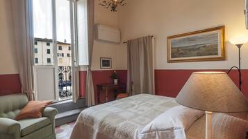 Une chambre d’hôtel avec un lit, une chaise, une table, une lampe, un tableau et une fenêtre avec des rideaux.