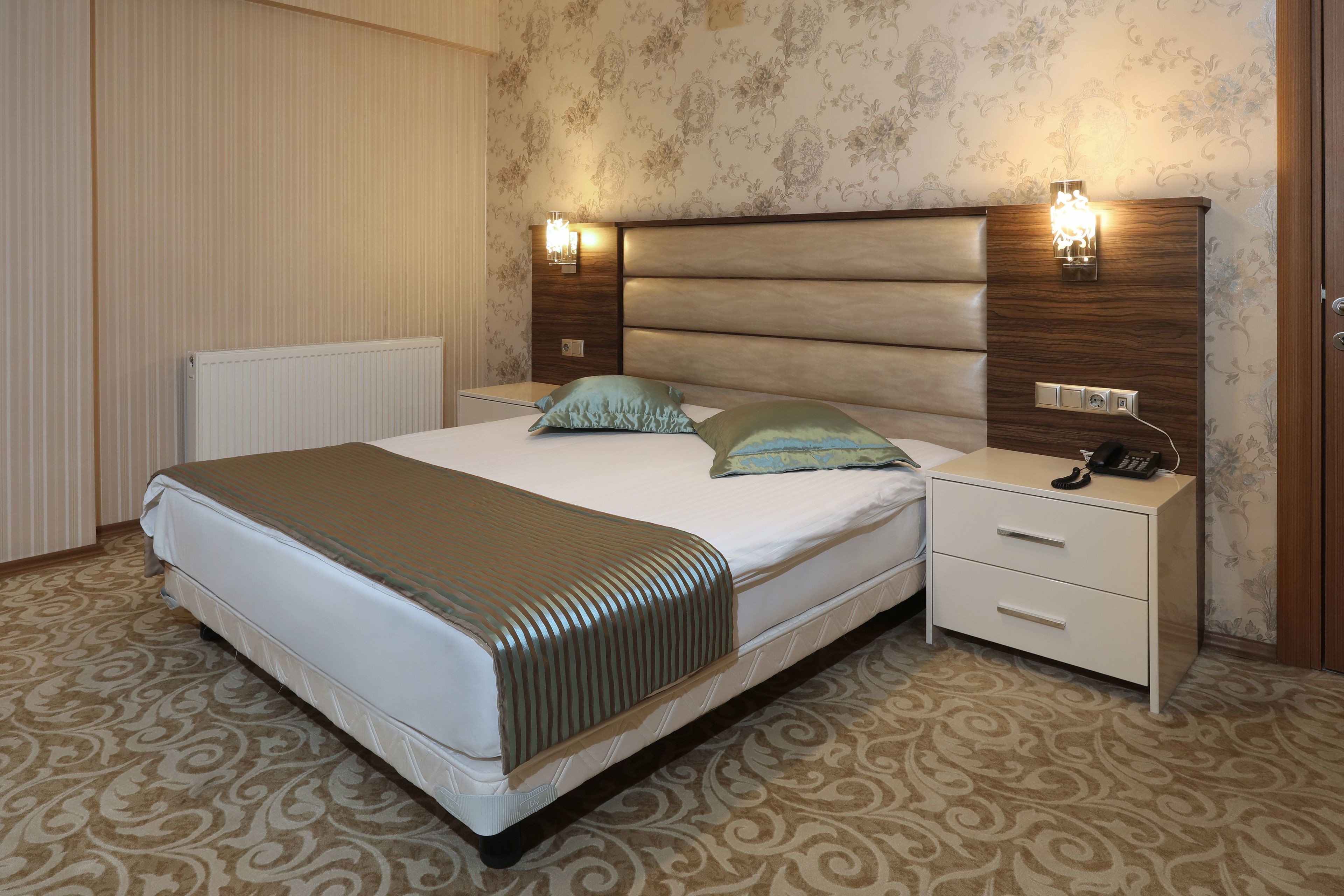 Una cama bien hecha con cabecera, dos mesitas de noche con lámparas y una alfombra estampada.