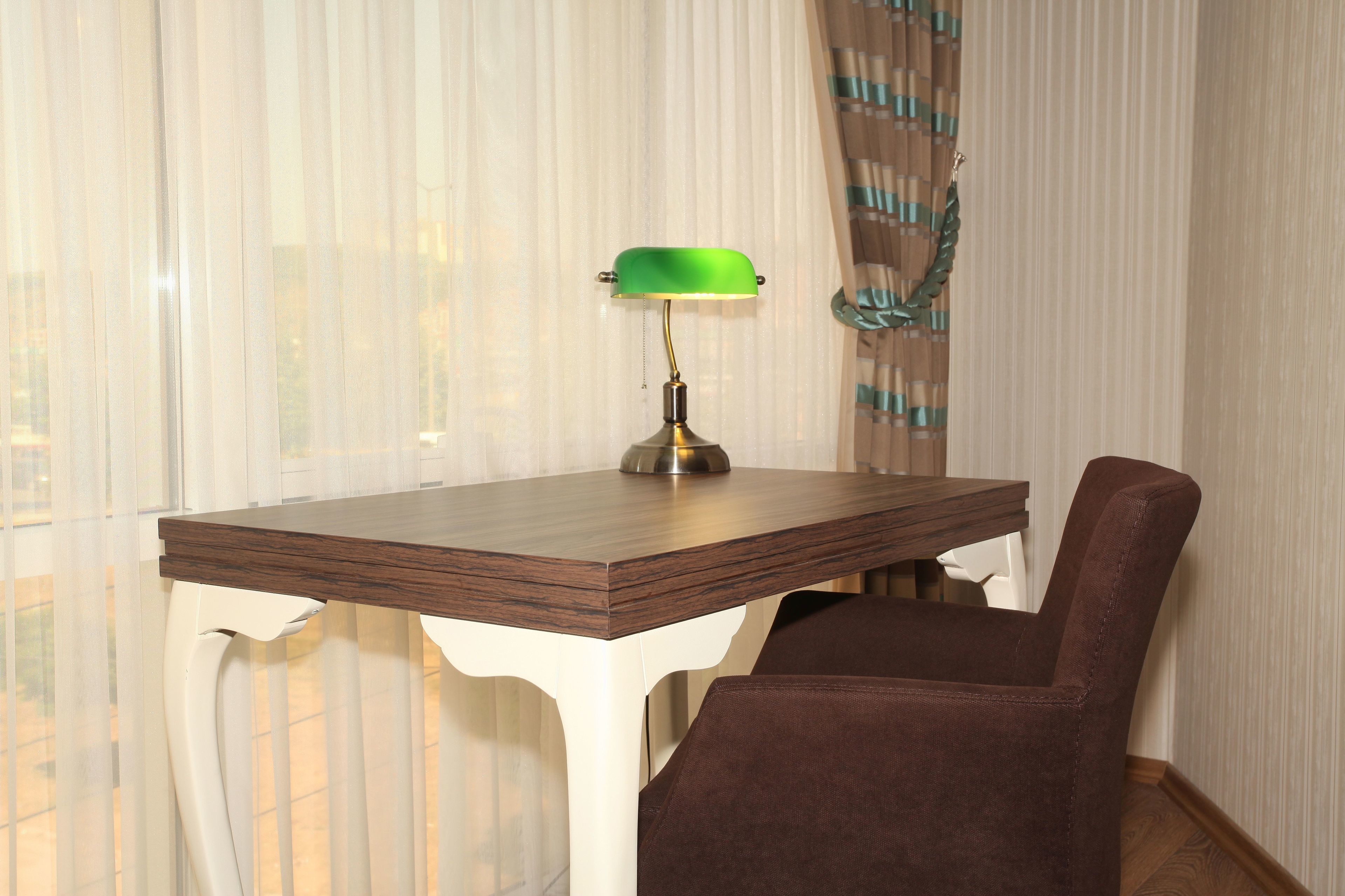 A dining table with a green lamp, a brown chair, and a window with sheer curtains.