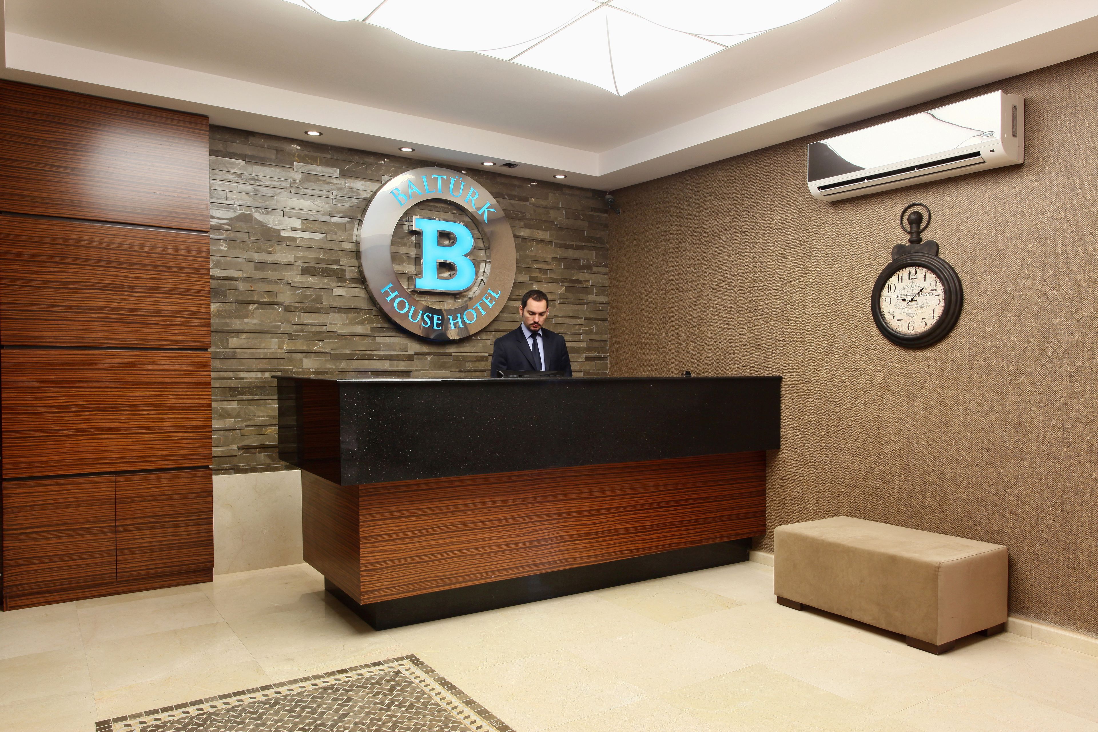 A reception area with a wooden reception desk, a stone wall with a 'BALTÜRK HOUSE HOTEL' sign, a clock, and a bench.
