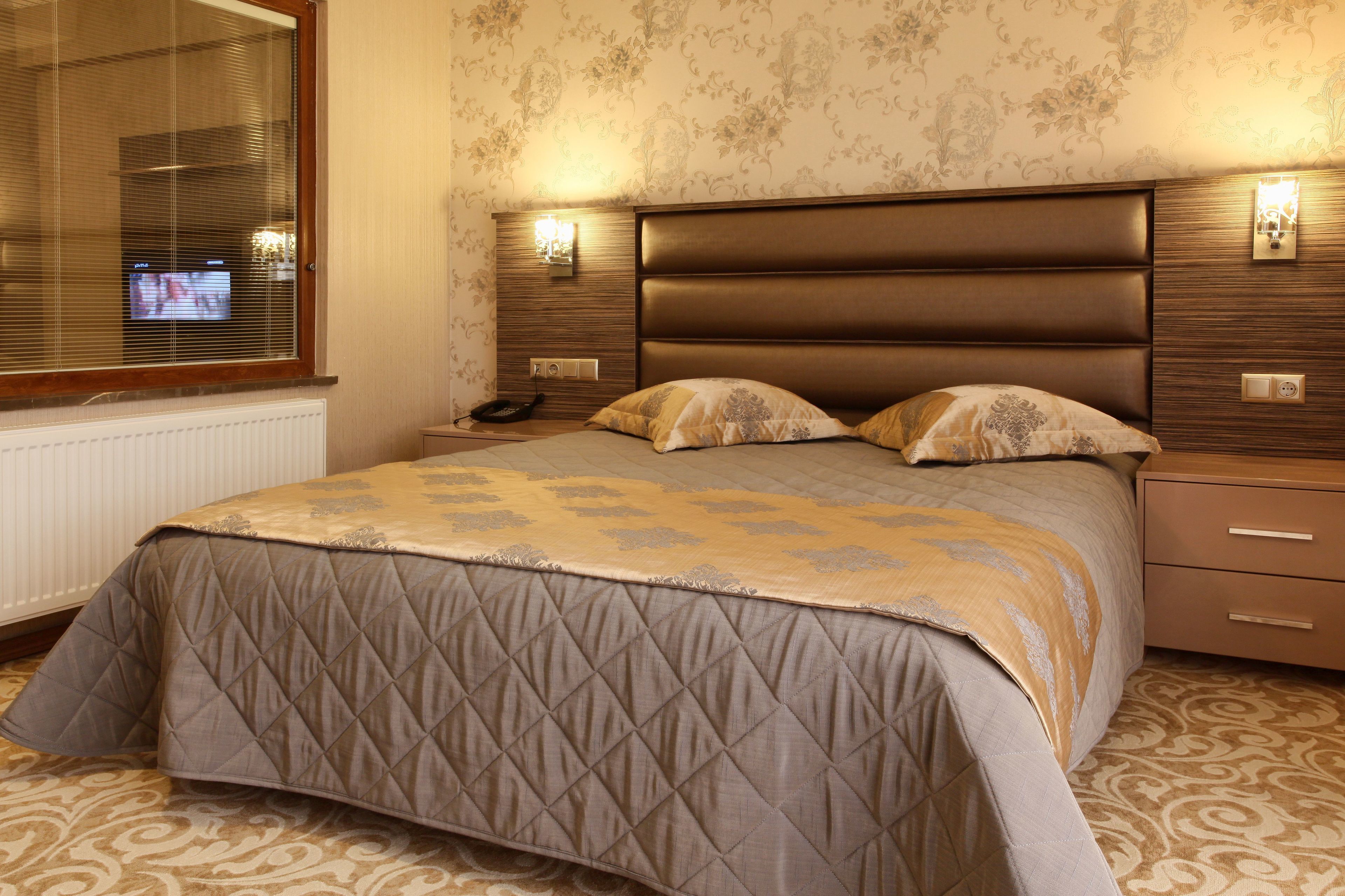 Un dormitorio con una cama grande, una mesita de noche, una lámpara de pared y un ventanal con persianas.