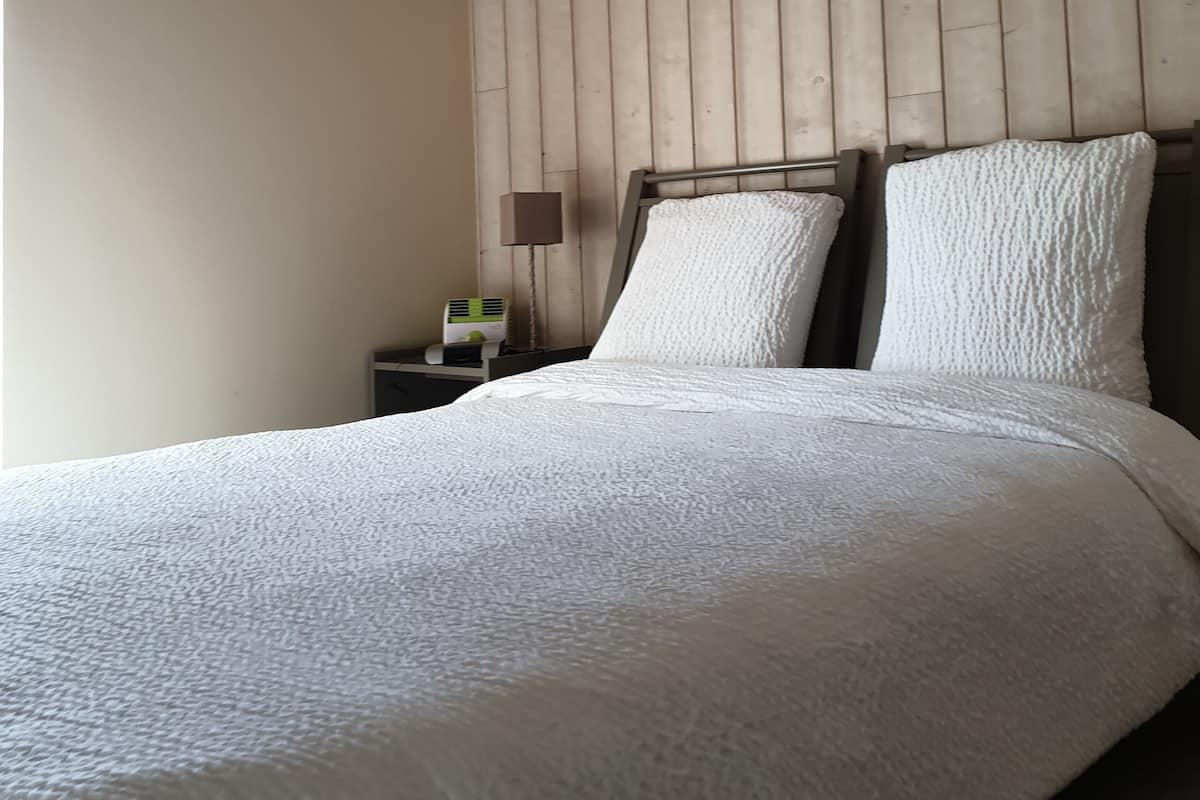 Une chambre avec un plafond en pente en bois, un lit avec une literie blanche, une table de chevet avec une lampe et une petite table avec une radio.