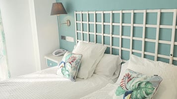 Una cama bien hecha con ropa blanca y almohadas florales, una mesita de noche con lámpara, y un aplique de pared.