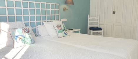 A neatly made bed with white bedding and decorative pillows, a bedside table with a lamp, and a chair with a folded towel.